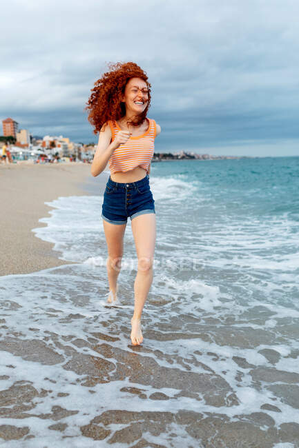 Full body of happy barefoot female traveler with flying long red hair running along sandy beach washed by foamy waves in windy weather — Stock Photo