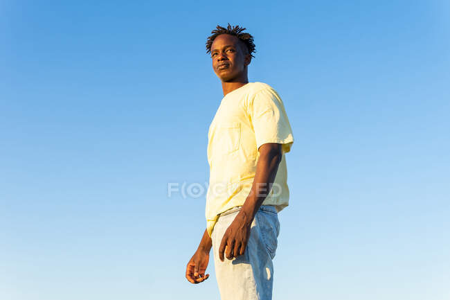 Baixo ângulo de jovem afro-americano pensativo em roupa da moda em pé contra o céu azul claro na noite de verão olhando para longe — Fotografia de Stock
