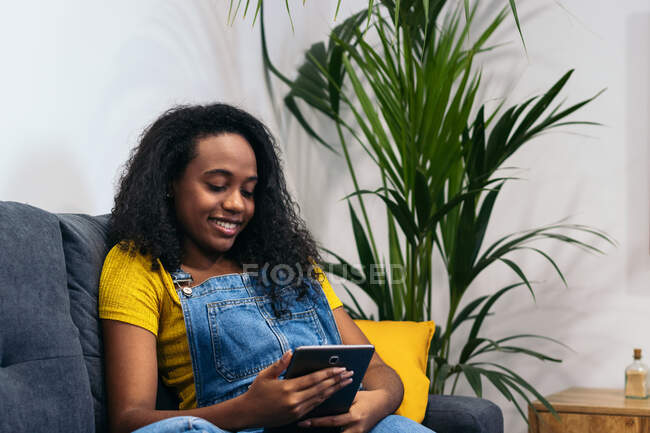 Mujer afroamericana positiva en traje de mezclilla sentada en el sofá y sonriendo mientras navega por la tableta en casa - foto de stock