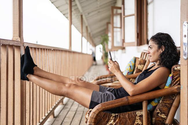 Vista lateral de la alegre joven turista en ropa casual sonriendo mientras está sentada usando un teléfono inteligente en un sillón en una terraza de madera de una casa envejecida en un día soleado - foto de stock