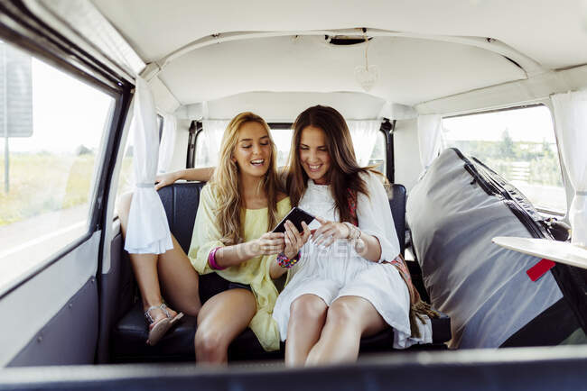 Dos chicas lindas sentadas dentro de una furgoneta vestida con ropa de verano sonriendo mientras miran la pantalla de un teléfono inteligente - foto de stock