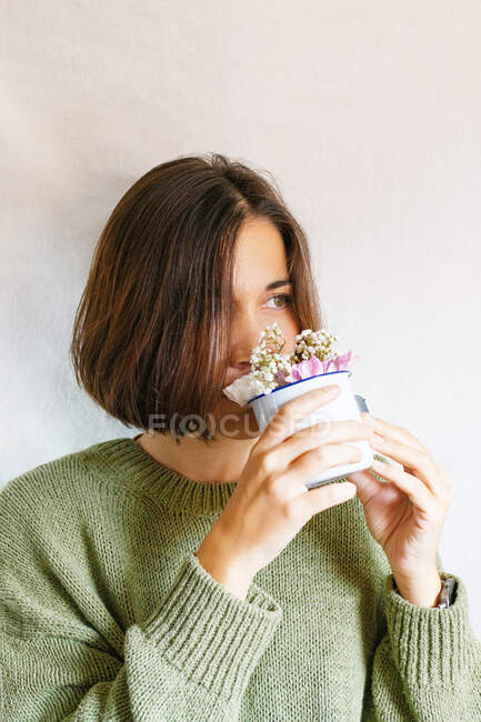 Joven hembra en jersey de punto oliendo flores sobre fondo gris - foto de stock