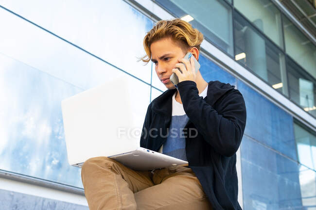 Focused young male freelancer in casual wear having phone conversation while sitting near modern building with netbook on street of city — Stock Photo