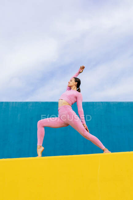 Corps entier jeune femme portant des vêtements de sport à la mode faisant Viparite Virabhadrasana pose sur un mur jaune et bleu sous un ciel nuageux — Photo de stock