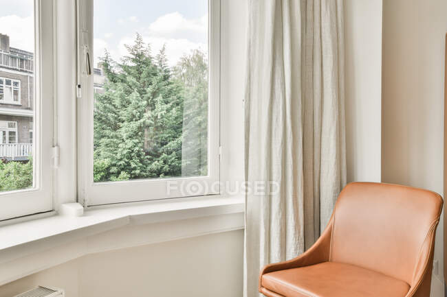 Потішний шкіряний стілець, розташований біля вікна й завіси у світлій кімнаті сучасного будинку. — стокове фото