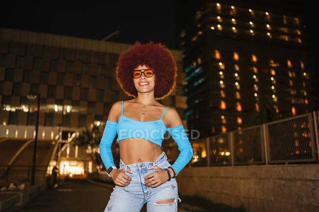 Positive Frau mit Afro-Frisur trägt trendige Klamotten und blickt in die Kamera, während sie nachts auf der Straße mit Gebäuden steht — Stockfoto