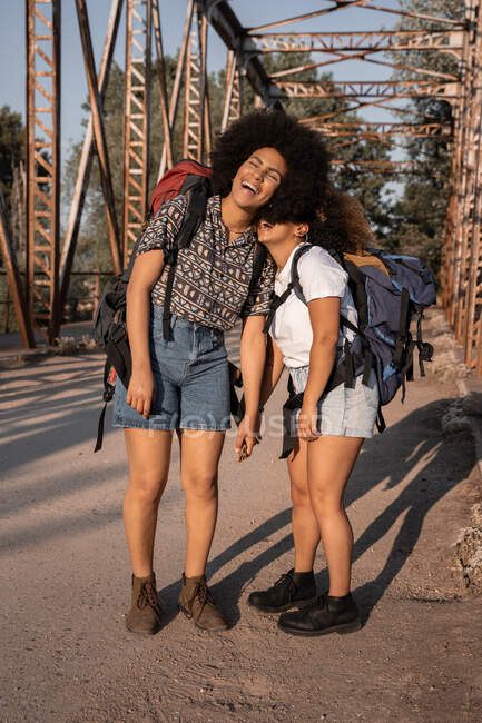 Des amies afro-américaines riantes avec des sacs à dos s'embrassant les unes les autres les yeux fermés sur un pont rouillé pendant une randonnée — Photo de stock