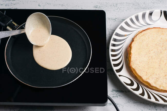 Сверху здорового теста для кето блинов, выливающихся из ковша на черную сковородку на плиту на светлой кухне — стоковое фото