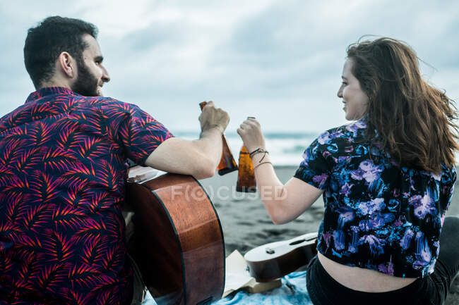 Back view of positive couple of musicians sitting with guitars and clinking bottles of beer while sitting on sandy beach near ocean in daytime — Stock Photo