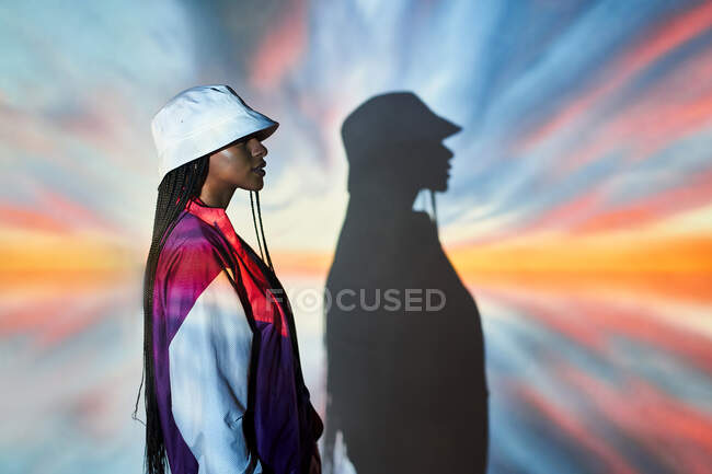Side view of self assured young Dominican female teenager in trendy outfit and hat standing near wall with creative abstract projections and looking away — Stock Photo