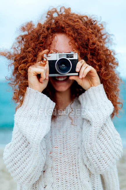 Positive ginger haired female in knitted sweater taking picture on retro photo camera on coast of sea — Stock Photo