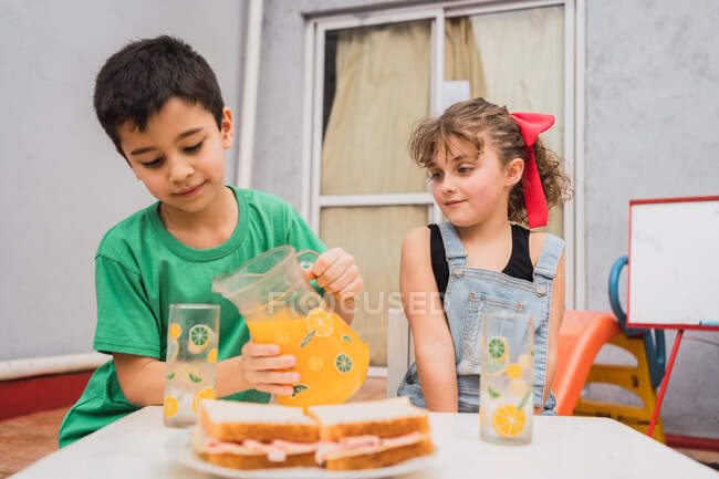 Menino derramando suco doce de jarro enquanto sentado perto da menina à mesa com sanduíches frescos na placa na sala de luz — Fotografia de Stock
