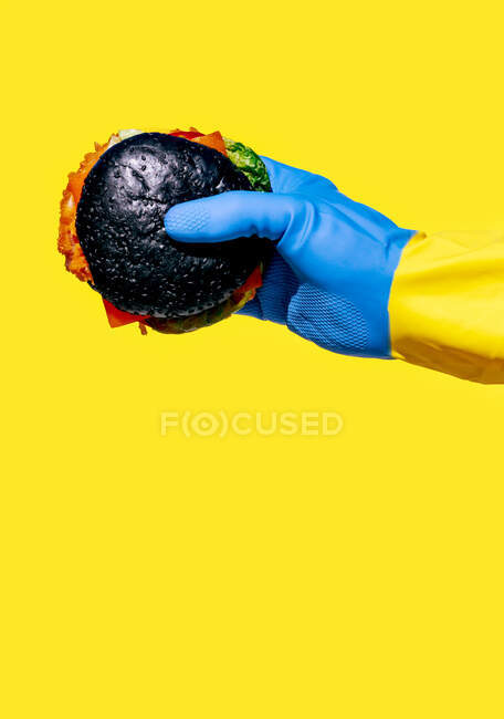 Crop person in colorful rubber glove demonstrating burger with black bun as unhealthy food concept against yellow background — Stock Photo