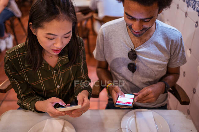 Счастливая молодая многорасовая пара в повседневной одежде с помощью смартфонов, сидя вместе за столом в современном ресторане — стоковое фото