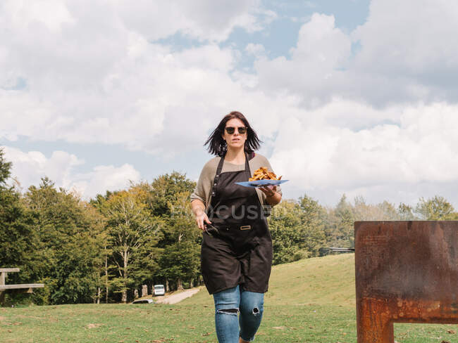 Серьёзная женщина-повар в фартуке несет тарелку с жареными куриными крылышками во время прогулки возле гриля на травянистом лугу в сельской местности — стоковое фото