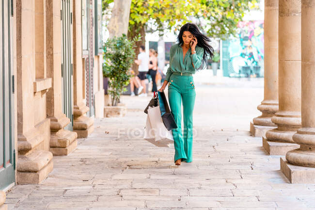 Mujer comprador serio en traje de moda con bolsas de compras que tienen conversación telefónica en el teléfono inteligente mientras pasea por la calle en la ciudad - foto de stock