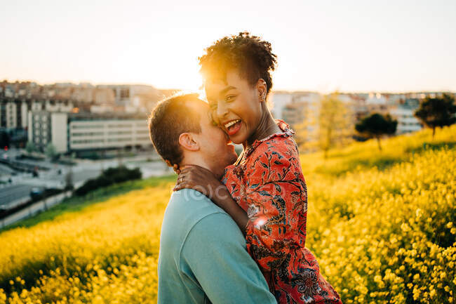 Visão lateral da jovem mulher afro-americana feliz com o cabelo curto sorrindo enquanto abraça o namorado com os olhos fechados em pé no prado florescendo no dia ensolarado — Fotografia de Stock
