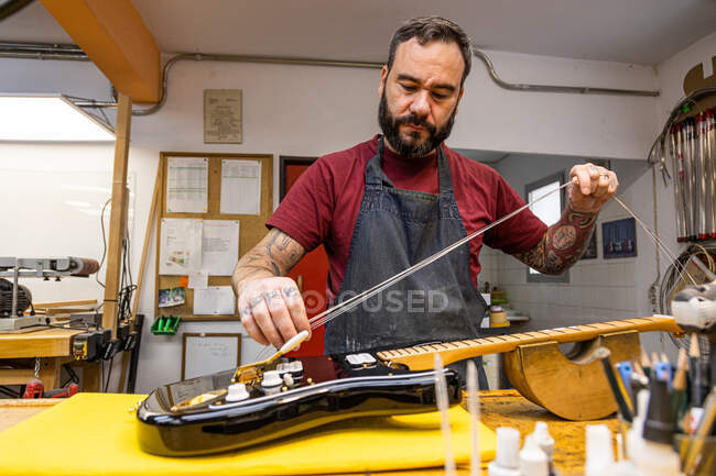 Abile artigiano in grembiule in piedi e cambiando corde sulla chitarra elettrica in studio di laboratorio professionale — Foto stock