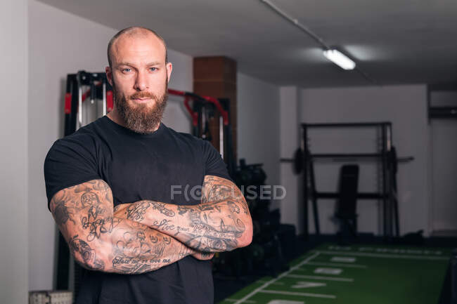 Strong adult sportsman with beard and tattoos on folded arms looking at camera in gymnasium — Stock Photo