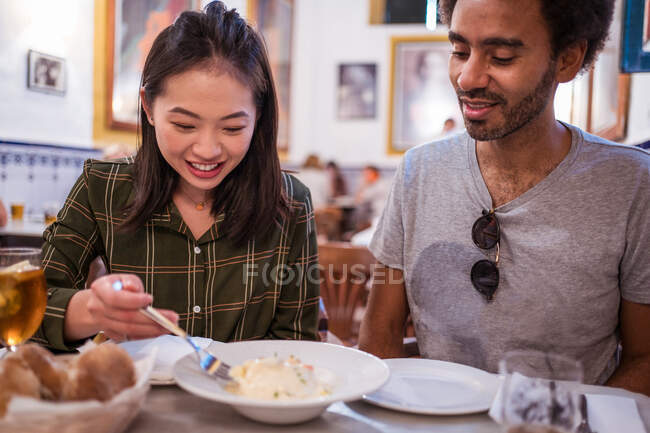 Jovem senhora asiática feliz com cabelo escuro em roupas casuais sorrindo enquanto come deliciosa salada durante o almoço com namorado étnico no restaurante — Fotografia de Stock