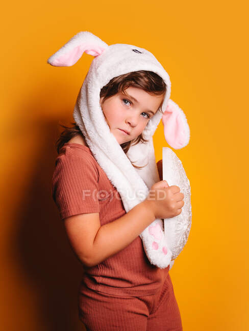 Vista lateral da menina triste com cabelo castanho em roupas casuais e chapéu branco com orelhas de coelho em pé e olhando para a câmera contra fundo amarelo no estúdio — Fotografia de Stock