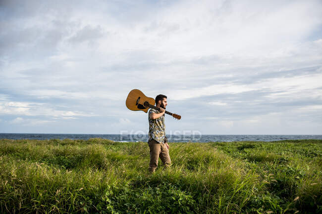 Спокойный человек музыкант в повседневной одежде, стоящий с акустической гитарой на плече среди зеленой травы на берегу океана летом при дневном свете — стоковое фото