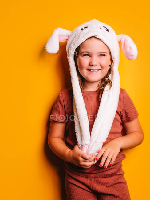 Feliz linda niña con cabello castaño y uñas arregladas con ropa casual y sombrero blanco con orejas de conejo de pie y mirando hacia otro lado contra el fondo amarillo en el estudio - foto de stock