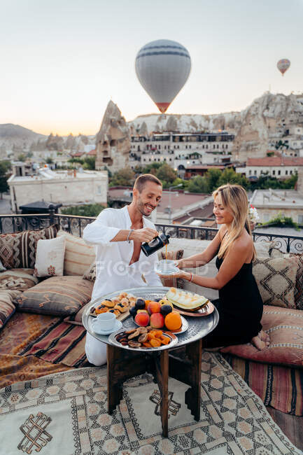 From above of cheerful male serving tea for wife while having romantic date on rooftop terrace against picturesque view of old city with hot air balloons flying — Stock Photo