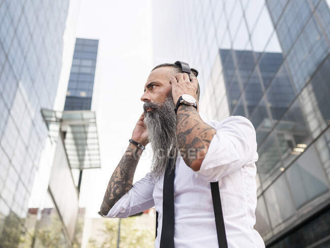Side view of serious bearded hipster male in formal wear listening to music with wireless headphones while standing near modern buildings — Stock Photo