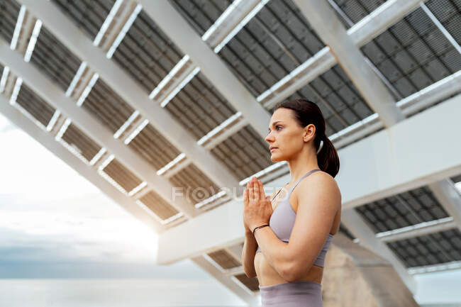 Side view of tranquil female in activewear doing namaste gesture with folded hands on street near solar panel — Stock Photo