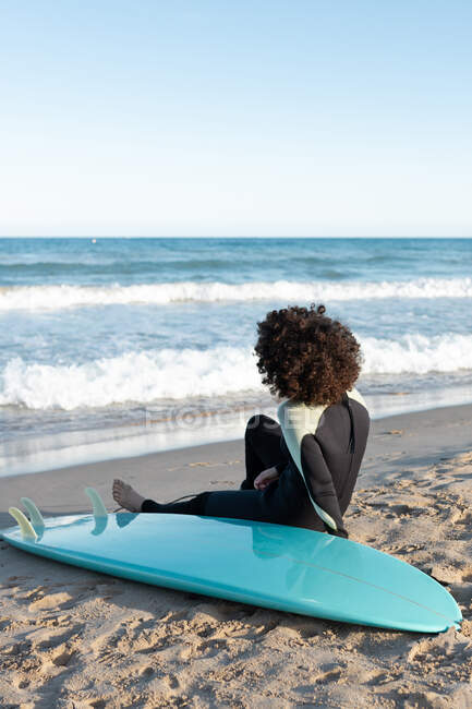 Voltar ver corpo inteiro de surfista fêmea descalça anônimo em wetsuit sentado na praia de areia perto do mar ondulando — Fotografia de Stock