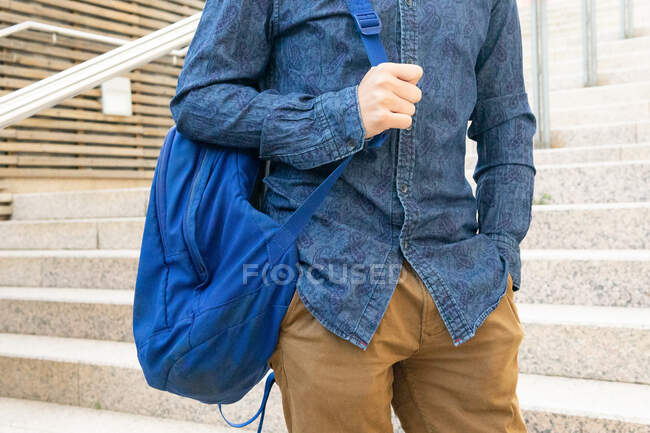 Crop anonymous male student in stylish outfit with blue rucksack standing near stone steps of building on street of city — Stock Photo