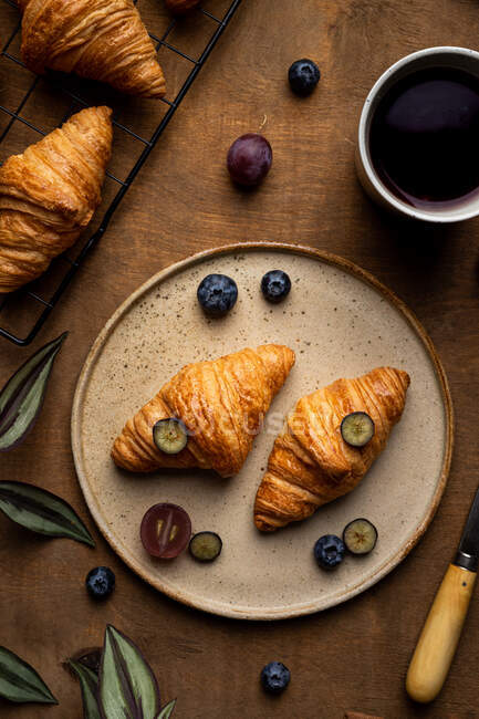 From above of tasty fresh baked croissants served on plate with fruits placed near cup of tea on wooden table in morning time in light room — Stock Photo