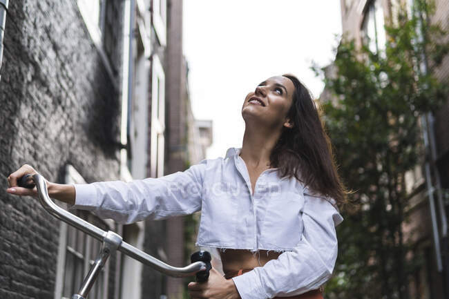 Lächelnde junge Frau in legerer Kleidung steht mit Fahrrad in der Nähe eines Gebäudes auf der Straße in der Innenstadt, während sie nach oben schaut — Stockfoto