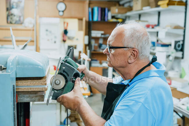Side view of concentrated senior male carpenter with white hair in apron and eyeglasses cutting wooden boards with circular saw during work in joinery — Stock Photo