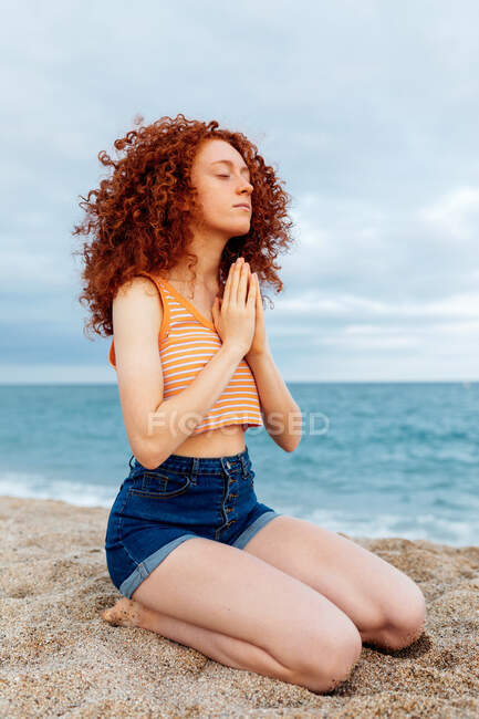Visão lateral corpo inteiro de mulher pacífica com cabelo encaracolado voando sentado de joelhos na praia arenosa e fazendo gesto namaste durante a meditação — Fotografia de Stock