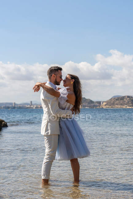 Side view of groom hugging bride while standing on shore near rippling sea during wedding celebration in nature on summer day — Stock Photo