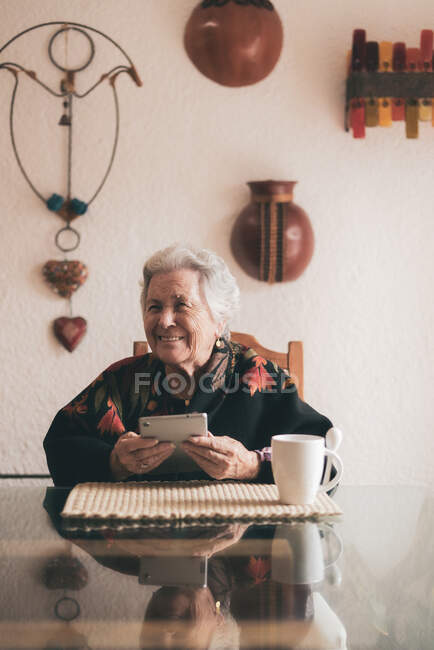 Улыбающаяся пожилая женщина в теплой одежде сидит за столом с табличкой и чашкой чая и смотрит в сторону — стоковое фото