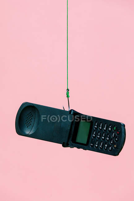 Старый черный открытый флип мобильный телефон висит на металлическом крючке с зеленой веревкой на розовом фоне в светлой современной студии — стоковое фото