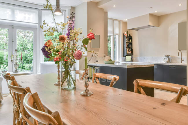 Salle à manger contemporaine avec bouquet floral fleuri dans un vase sur table en bois contre cuisine et fenêtres à la maison — Photo de stock
