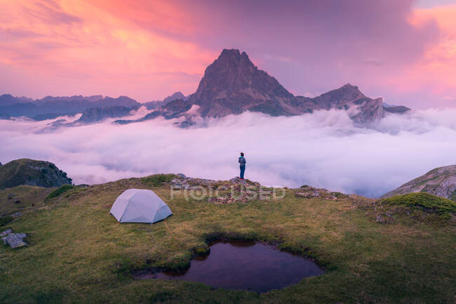 Далекий анонимный турист, стоящий на краю травянистой скалы с палаткой для кемпинга у горного хребта и густым туманом в вечернее время — стоковое фото