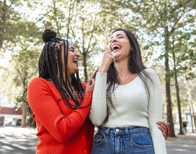 Glückliche multirassische Freundinnen in stylischer Kleidung stehen und lachen bei Tageslicht an grünen Bäumen auf der Straße — Stockfoto