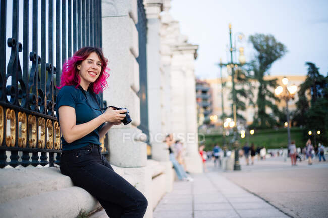 Позитивная женщина-фотограф с розовыми волосами и фотокамерой в руке, смотрящая в камеру, сидя рядом с забором в старом здании на площади — стоковое фото