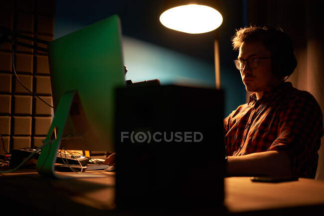 Концентрированный мужчина в клетчатой рубашке и очках, работающий за компьютером, сидя за столом с лампой и микрофоном во время записи подкаста — стоковое фото