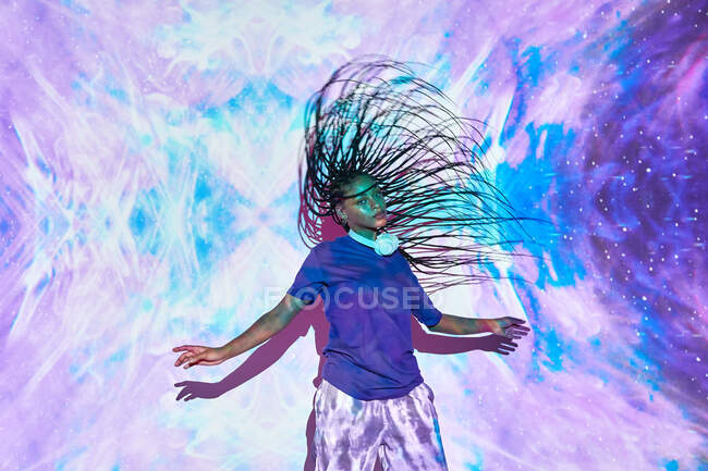 Side view of ethnic female millennial shaking long braided hair while dancing near wall with bright blue and lilac illumination — Stock Photo