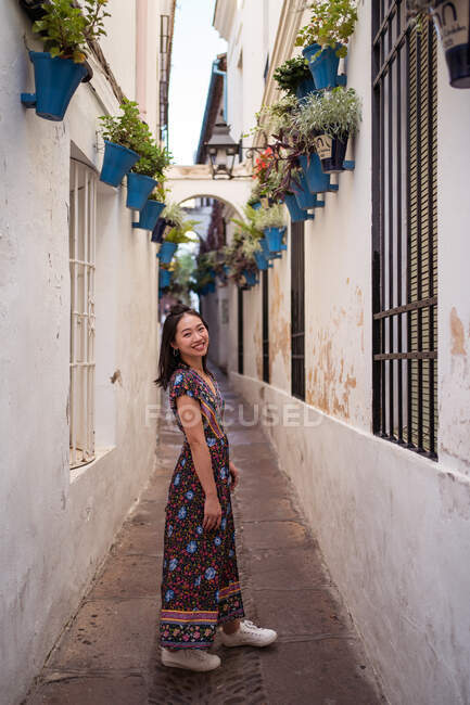 Full body vista laterale di sorridente asiatica turista in piedi in stretto passaggio tra le case, mentre guardando la fotocamera in città — Foto stock
