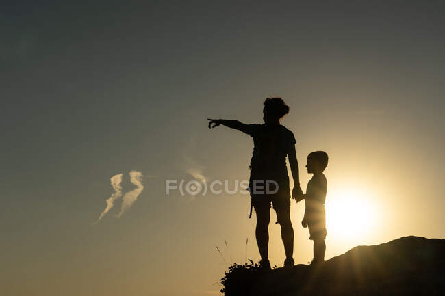 Madre e figlio contro la luce sagomata dal sole che punta con le braccia verso l'orizzonte al tramonto — Foto stock