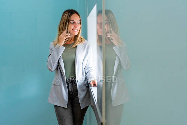 Mujer positiva en traje elegante hablando en el teléfono inteligente mientras mira por la ventana cerca de la pared en la sala de luz del edificio moderno - foto de stock