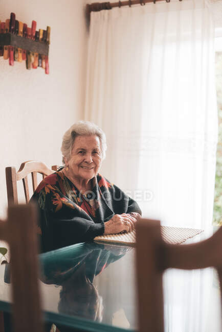 Mujer anciana con el pelo gris sentada en la mesa en la habitación con gran ventana cubierta de tul y sonriente mientras mira a la cámara a la luz del día - foto de stock