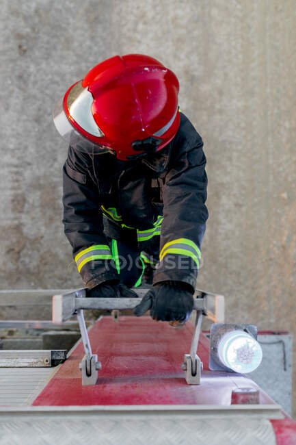 From above an anonymous firefighter wearing red protective helmet and uniform standing on fire truck ladder and looking away — Stock Photo